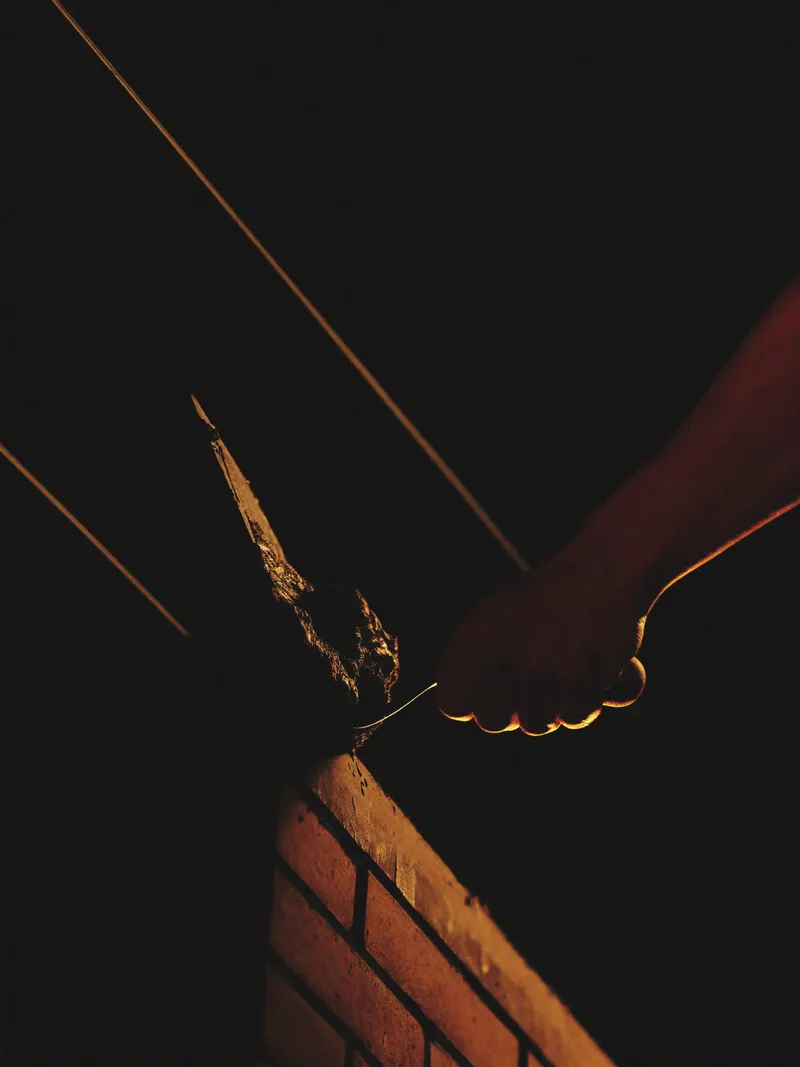 Hands buttering mortar onto a brick with a trowel above a wall course, lit by a single orange light