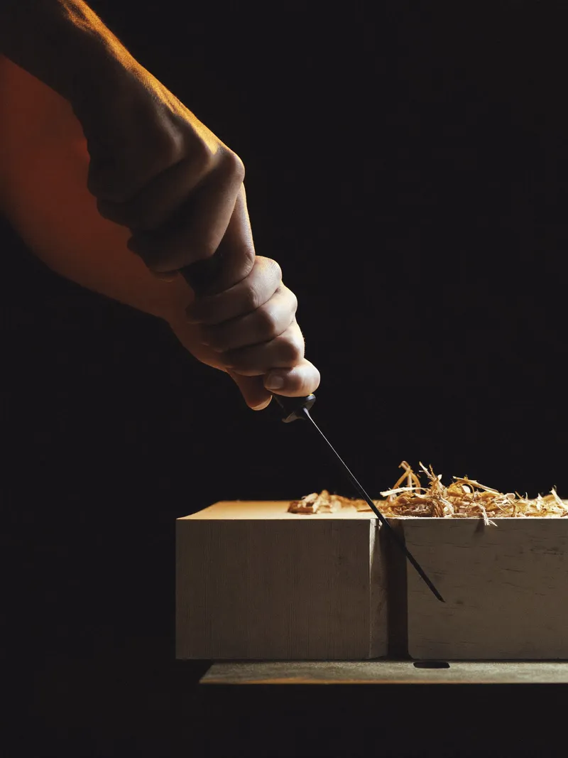 Hands guiding a chisel along a timber joint, lit by a single orange light