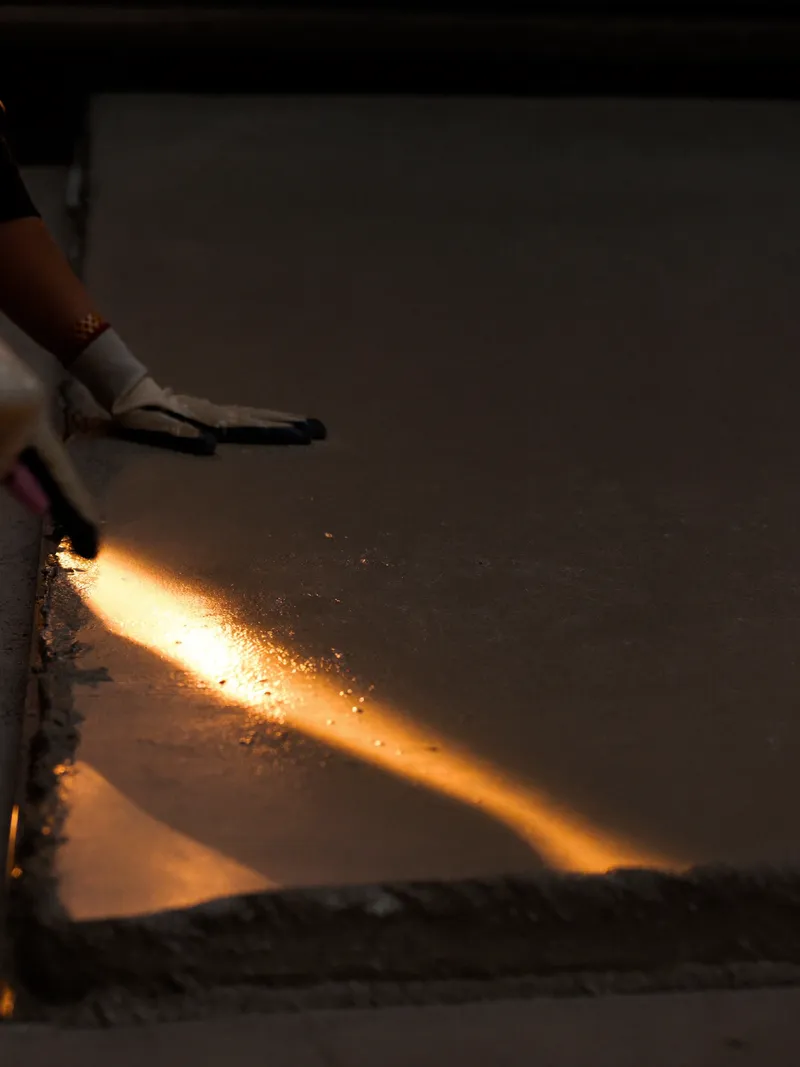 Gloved hands smoothing fresh wet concrete at the edge of a slab, lit by a single warm orange light