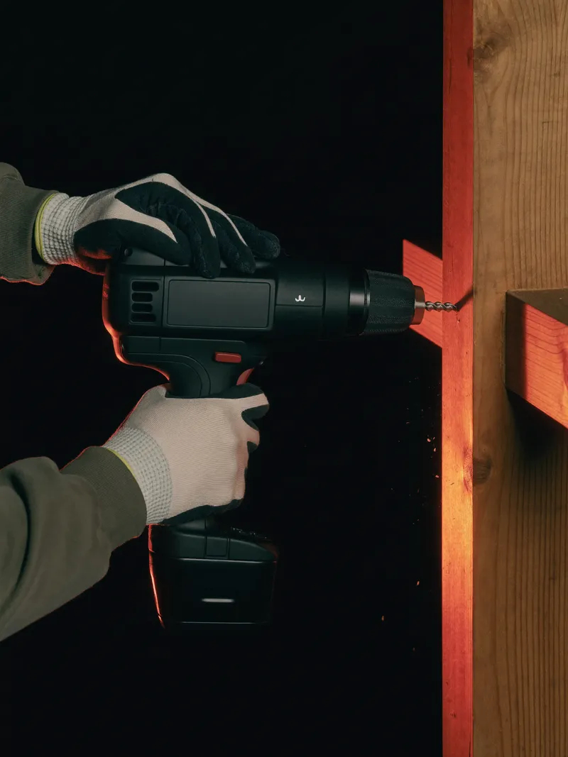 Gloved hands driving a screw through a timber fence rail with an impact driver, lit by a single orange light