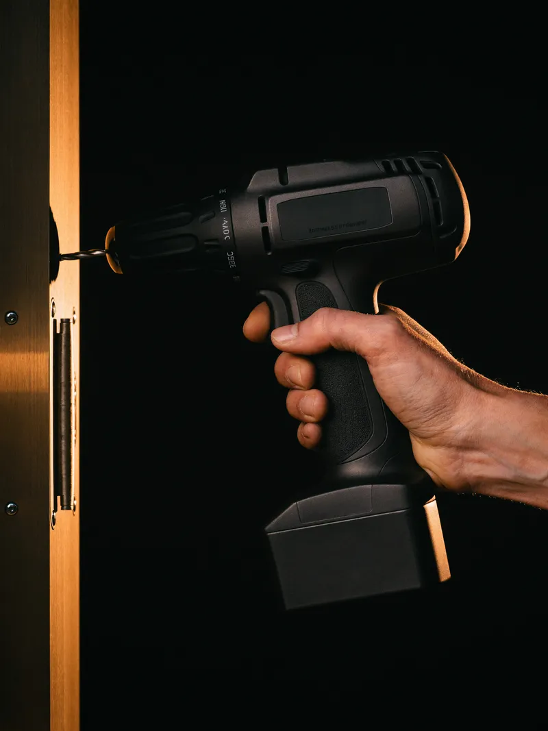 Hands driving a screw into a door hinge with a cordless drill, lit by a single orange light