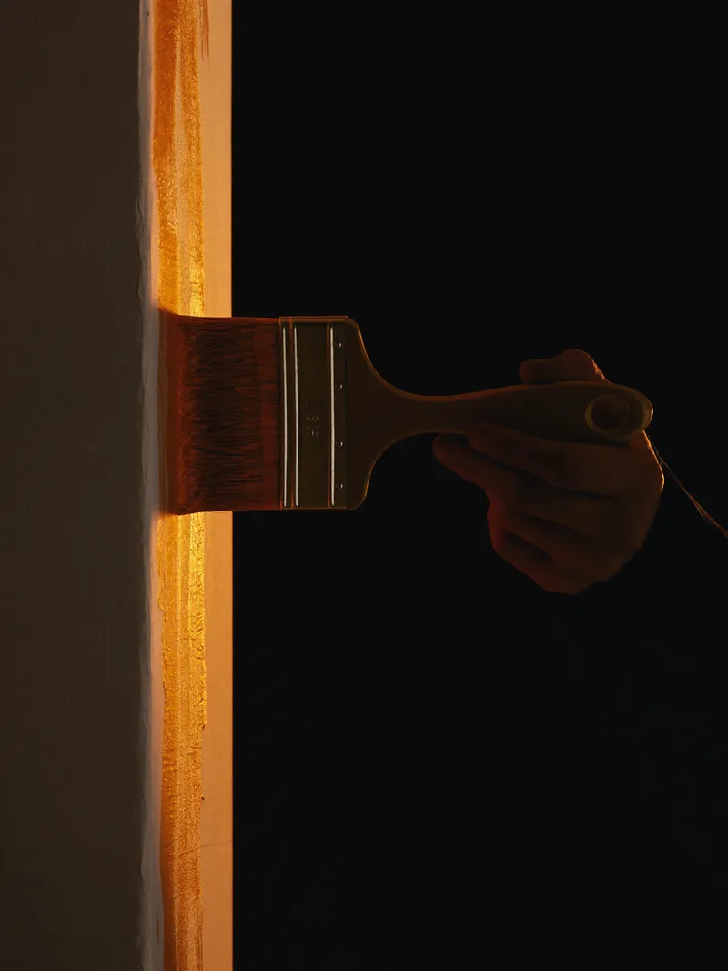 A painter cutting in a crisp edge with an angled sash brush, lit by a single orange light