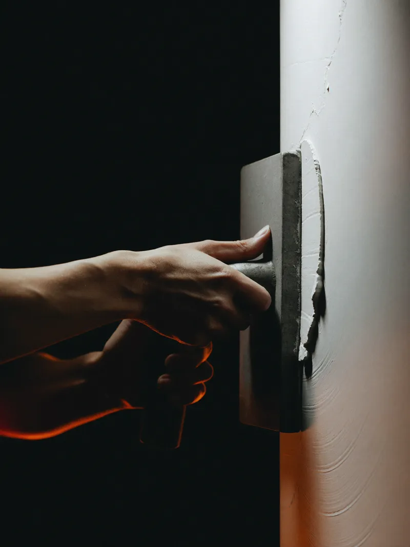 Hands smoothing fresh plaster on a wall with a steel trowel, lit by a single orange light