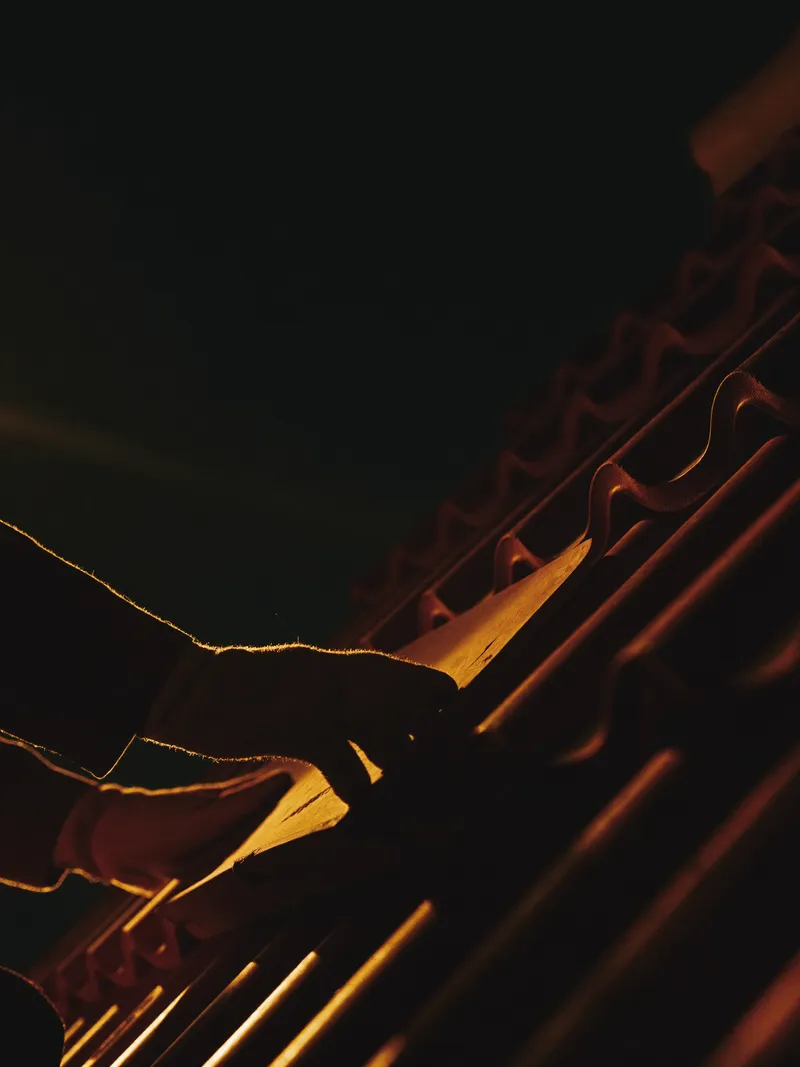 Work-gloved hands seating a terracotta roof tile, lit by a single orange light