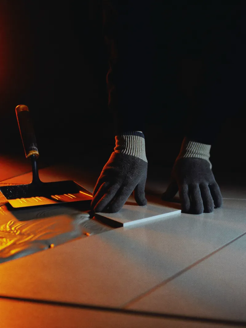 Gloved hands pressing a porcelain floor tile into adhesive, lit by a single orange light